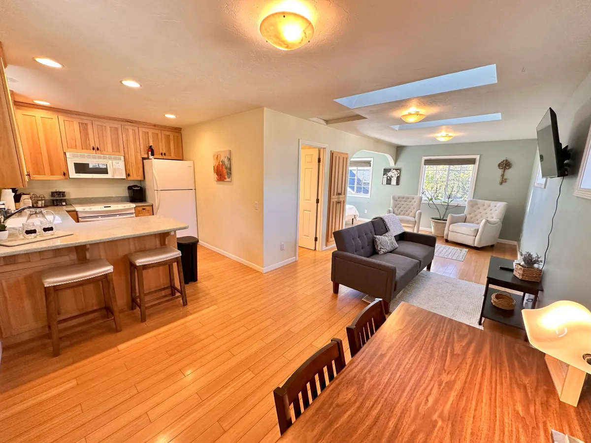 An open-plan interior that connects the kitchen area, dining table, and living area with a sofa. Natural light floods the space from large windows, creating a welcoming environment perfect for both relaxation and socialization.