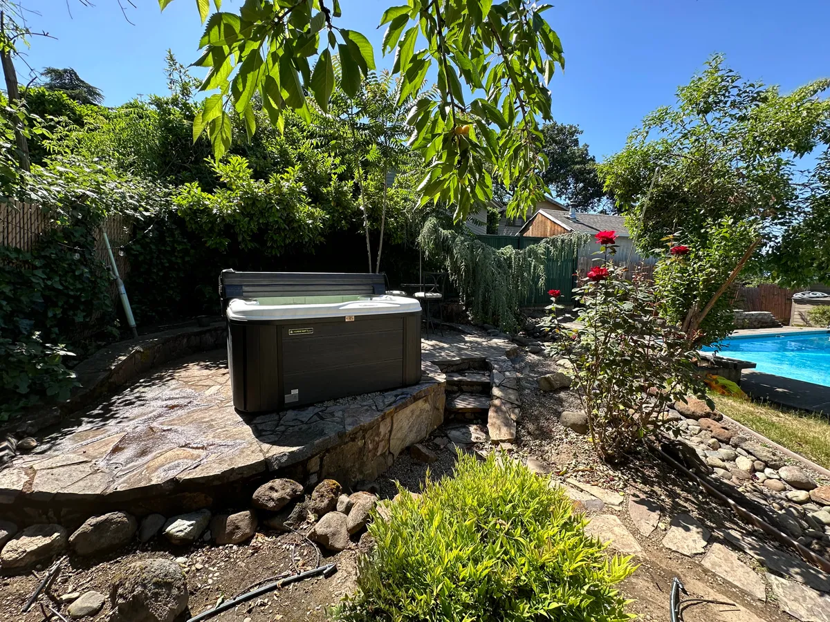 A serene outdoor space featuring a hot tub surrounded by flowering plants and a stone pathway, with a swimming pool nearby. The setting is tranquil, ideal for relaxing soaks or quiet swims while immersed in nature.