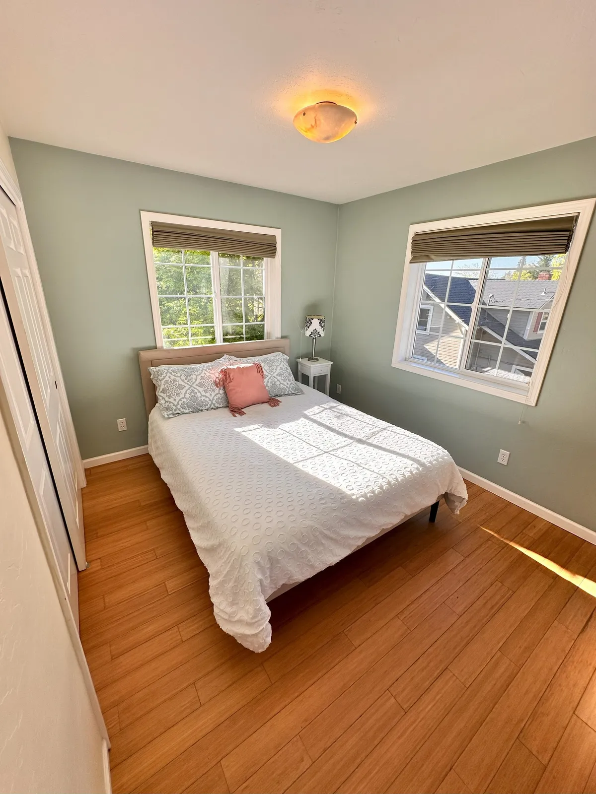 A serene bedroom scene featuring a plush bed dressed in white bedding and pillows, flanked by large windows that flood the space with natural light. The simple yet elegant decor creates a calming atmosphere ideal for rest and relaxation.