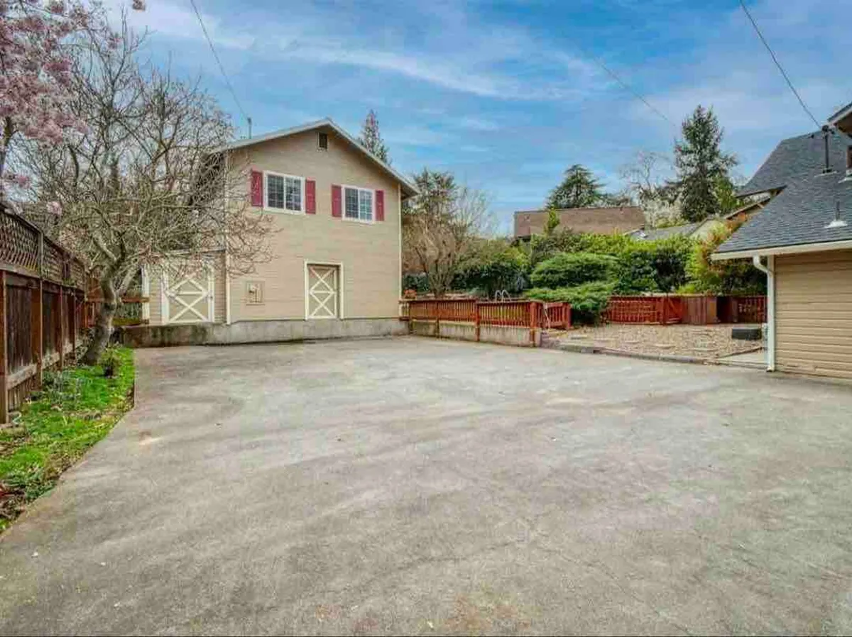 A two-story building with charming red shutters sits behind a fence, bordered by a paved driveway and surrounded by lush greenery. The scene feels inviting and grounded within a friendly, established neighborhood.