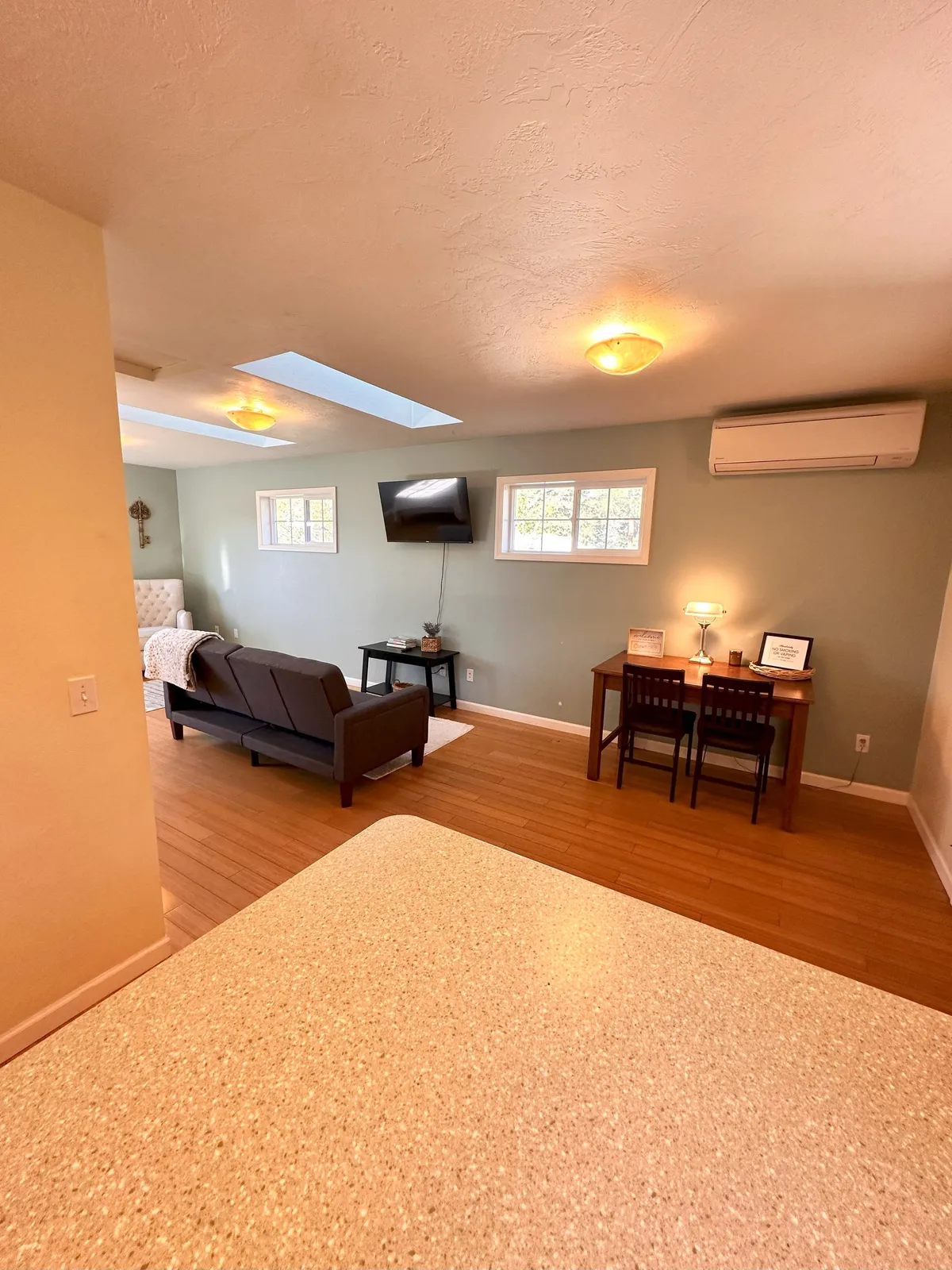 A cozy living area with a plush sofa, dining table, and a wall-mounted TV. Skylights bathe the space in morning sunlight, creating a bright and inviting atmosphere perfect for relaxing or entertaining. The seating arrangement and decor foster comfort and calm.