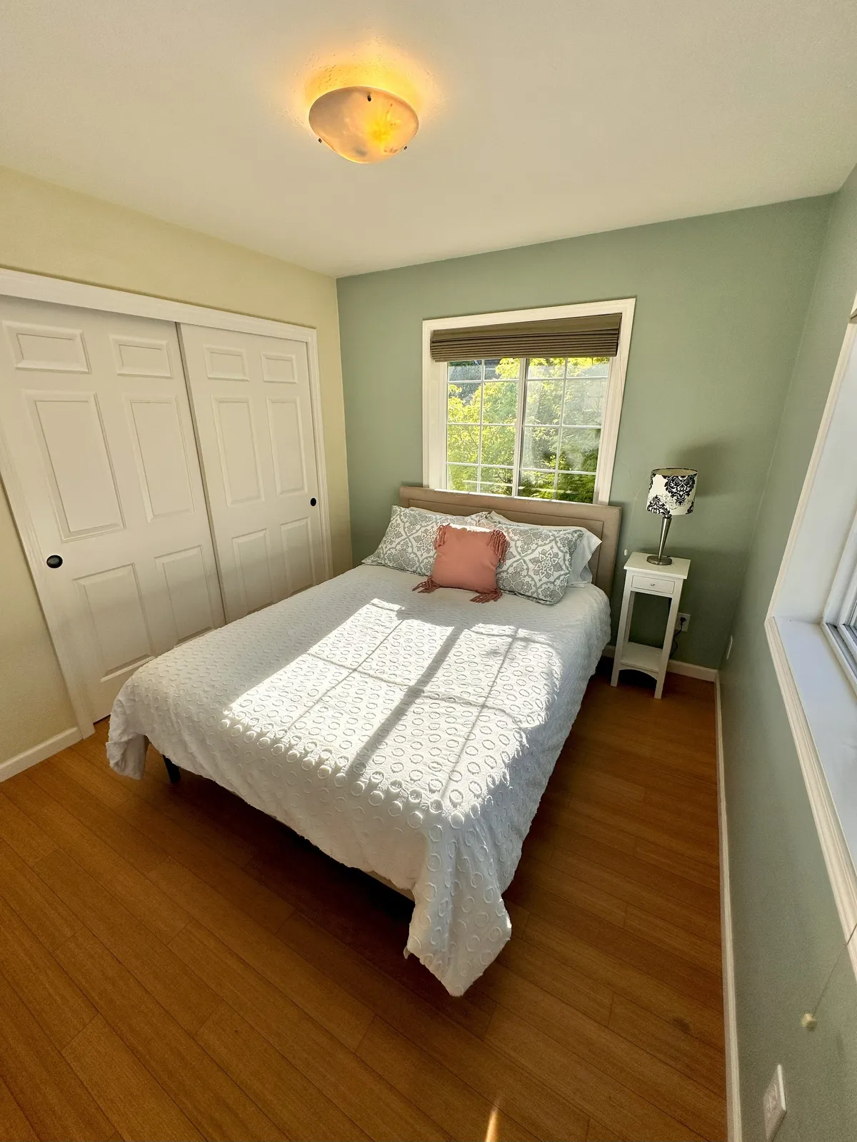 A cozy bedroom with a well-made bed, a window beside it, and a decorative lamp that provides a warm glow. The space feels intimate and perfect for unwinding at any time of day.