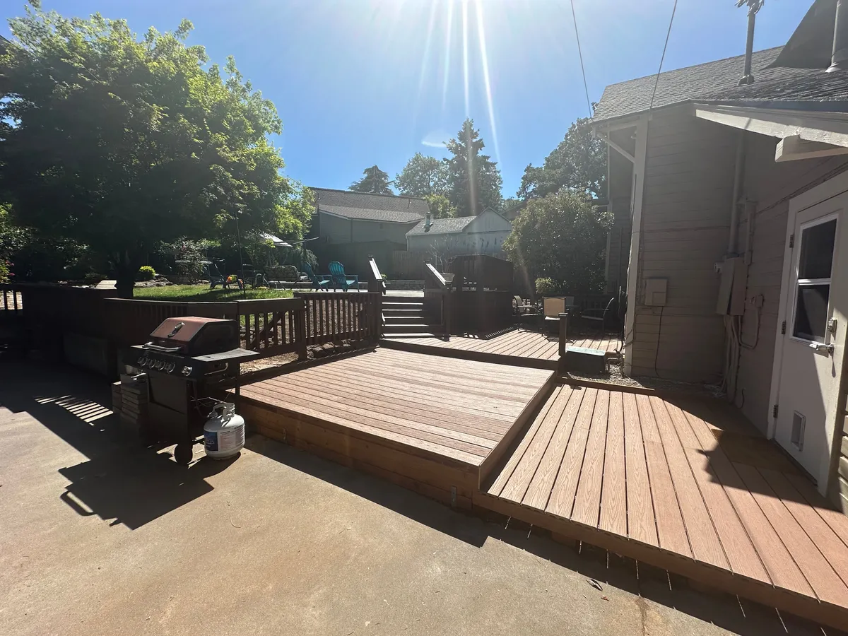 A sunny deck area features a grill, outdoor seating, and is framed by lush greenery, offering an inviting spot for outdoor dining or quiet reflection.