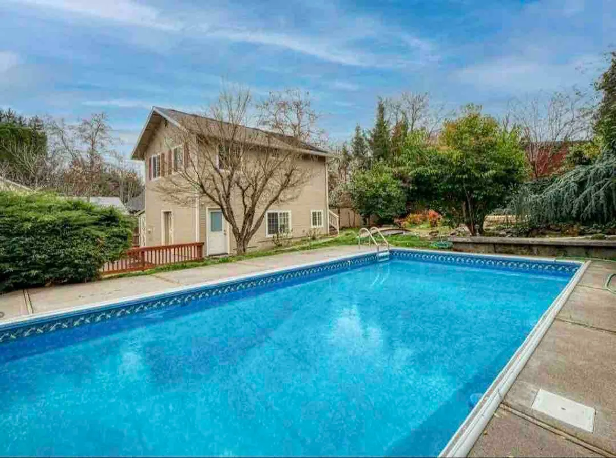 A serene exterior view featuring a sparkling swimming pool surrounded by lush greenery. The house sits in the background, and inviting poolside seating creates a relaxed, welcoming atmosphere. The scene hints at both quiet mornings and pleasant evenings by the water.